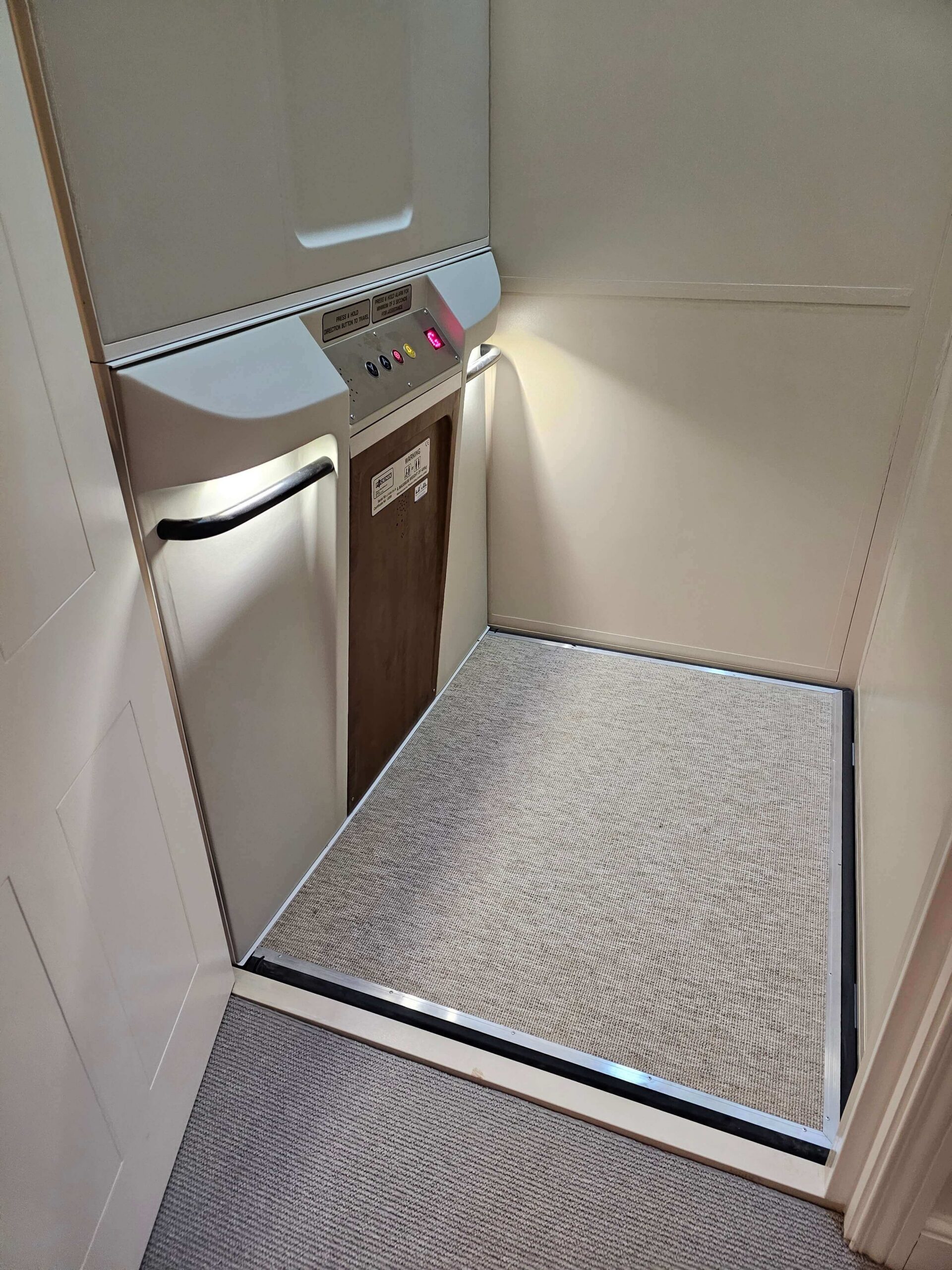 Interior view of a British Style vertical platform lift showing grey carpet flooring, timber inner door and control panel