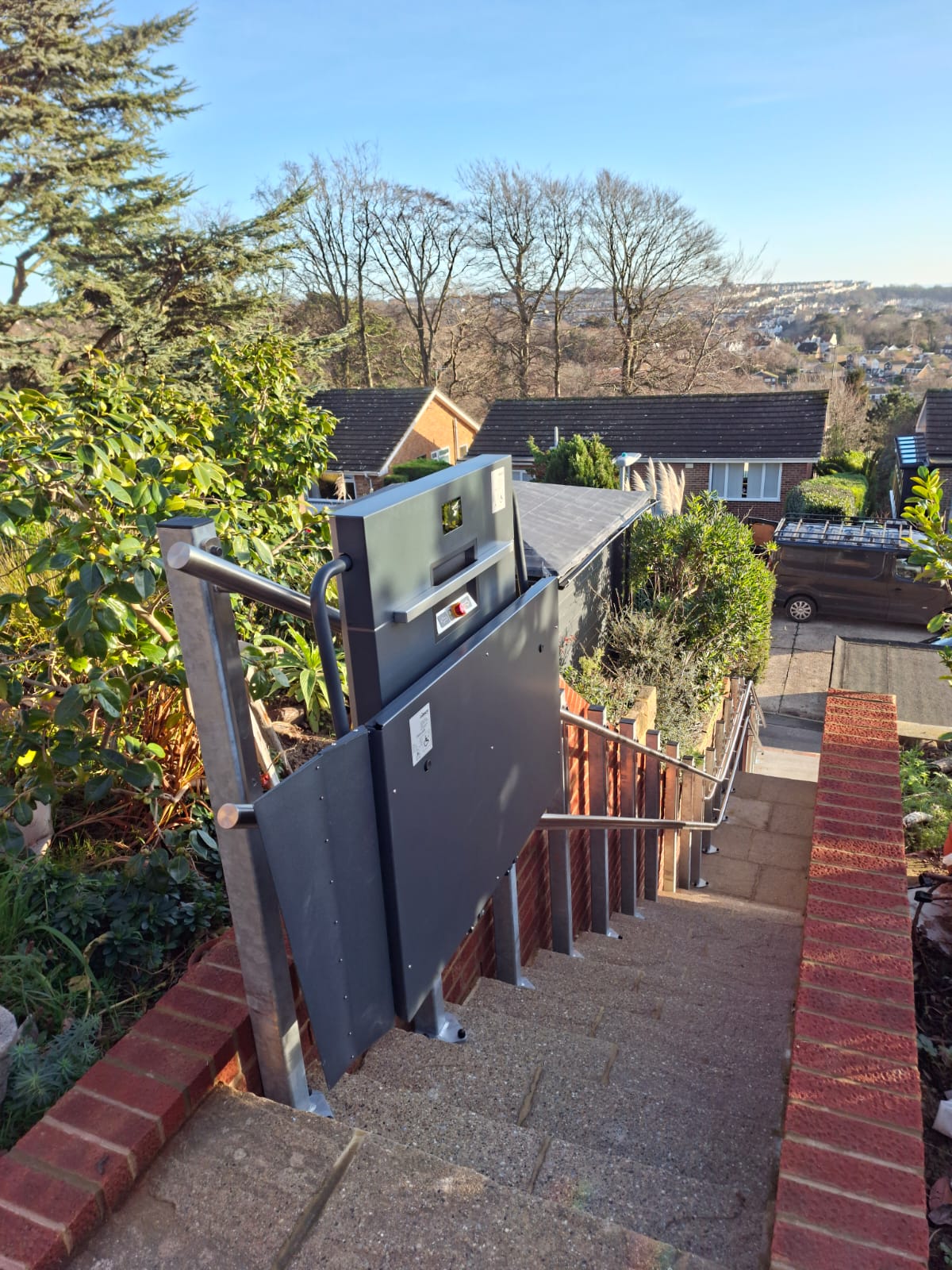 Grey inclined platform stair lift in folded upright position alongside a long flight of outdoor steps at a residential coastal property