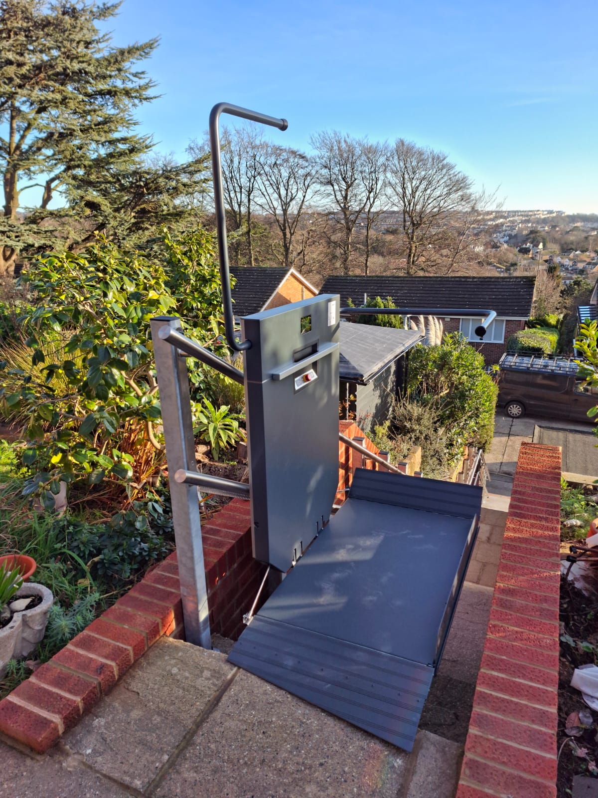 Grey inclined platform stair lift with boarding platform extended at the base of a steep flight of outdoor steps at a coastal residential property
