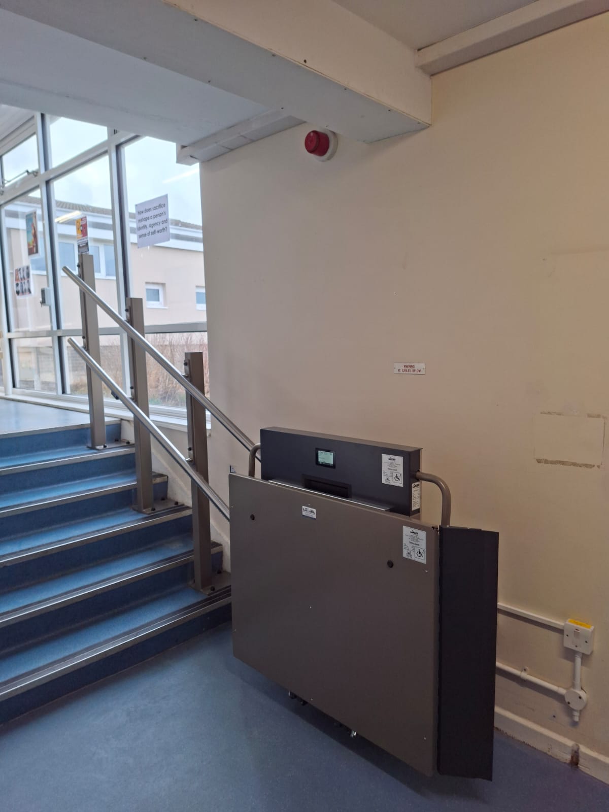 Compact platform stair lift installed beside blue-carpeted steps at Filey Academy, folded away when not in use.