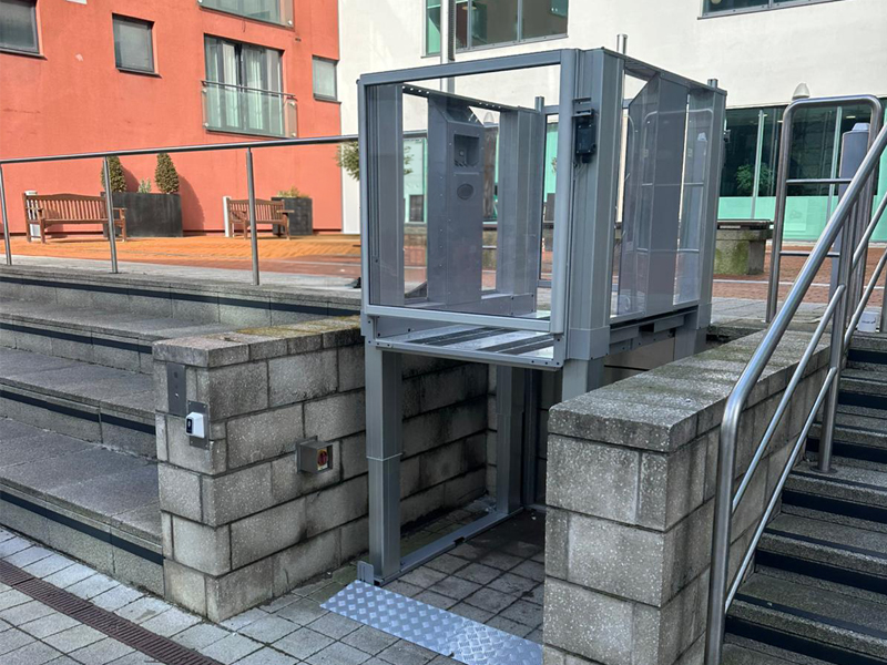 Tech Lift M4 external step lift at upper terrace level at Colton Square, Leicester, with glazed doors open and stainless steel handrails either side