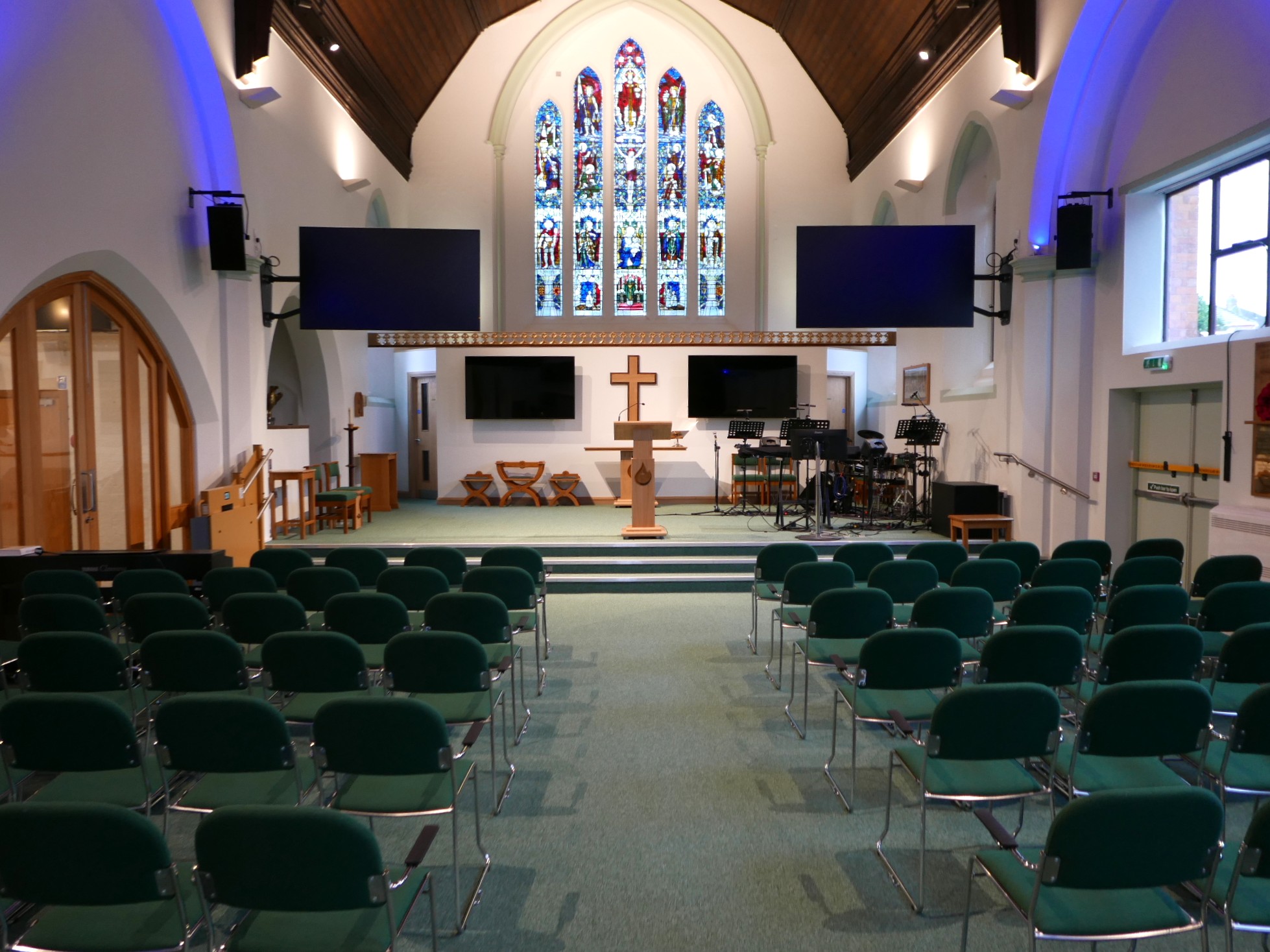 Interior of St Oswald's Church, Rugby, showing the nave with green seating, stained glass window and raised chancel area with a platform stairlift