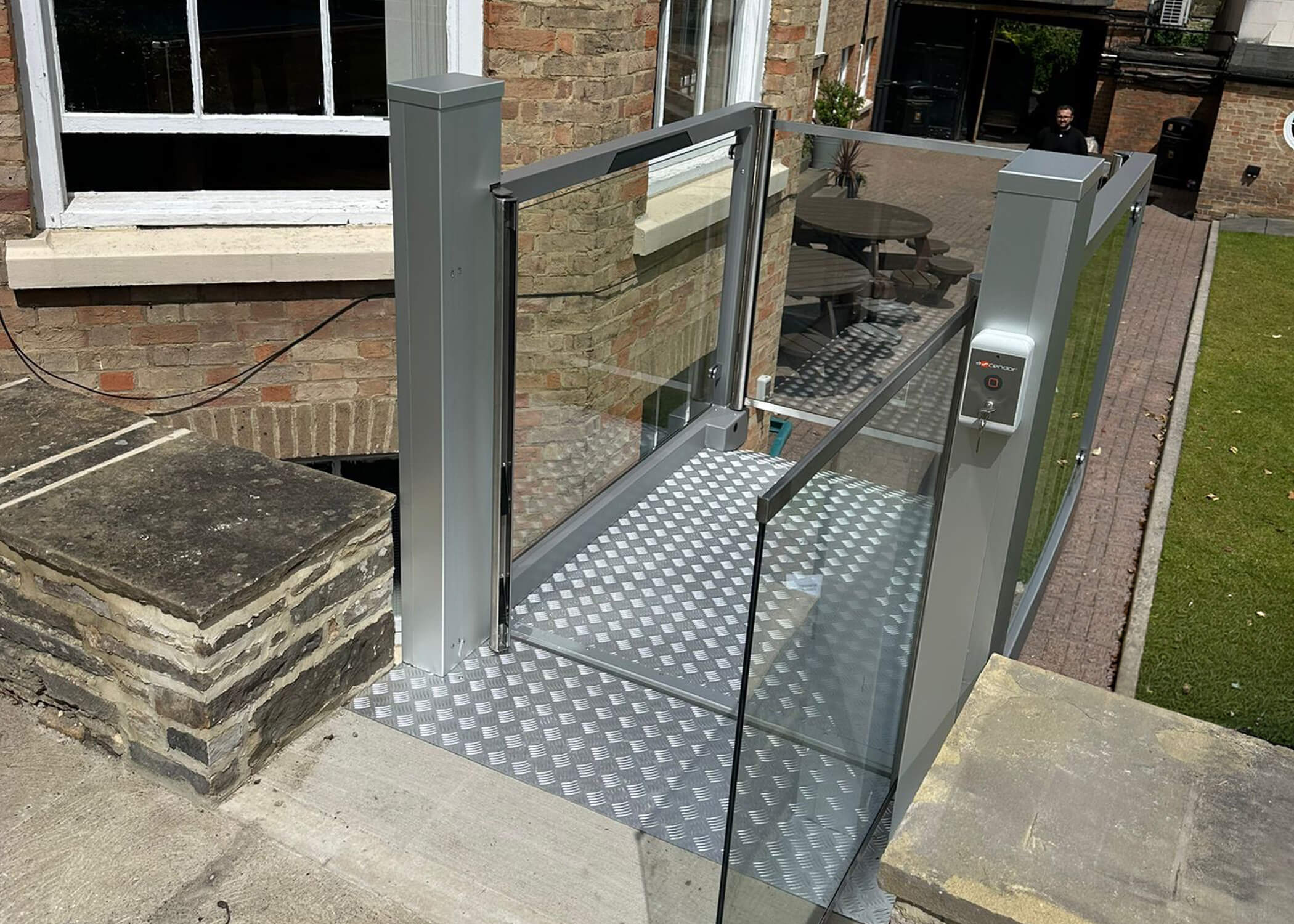 Glass-sided UnaPorte platform lift installed against a Grade II listed brick building at St Chad's College, with chequer plate platform and keypad control panel