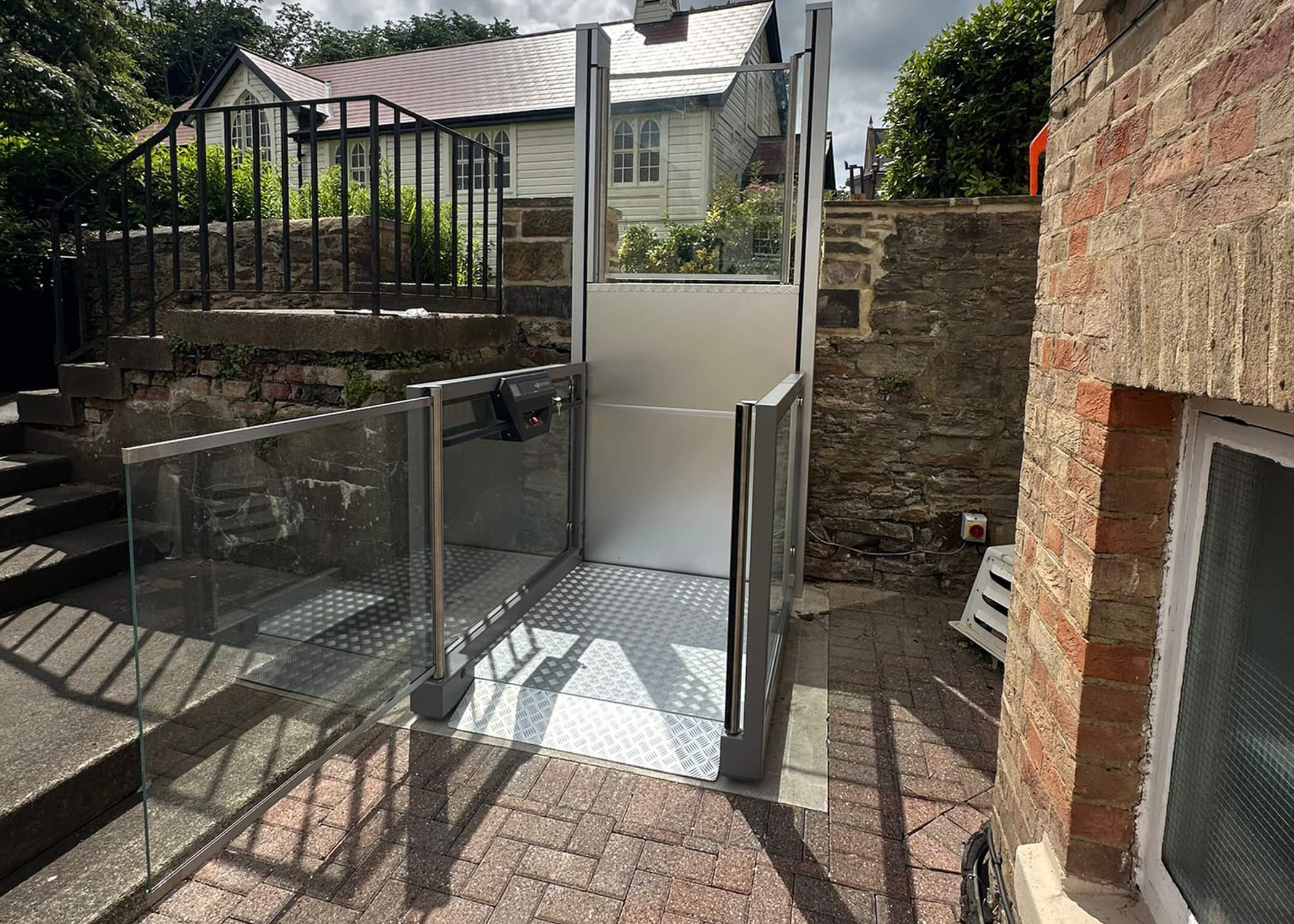 UnaPorte outdoor platform lift at ground level beside a stone wall at St Chad's College, with glass panels and chequer plate platform